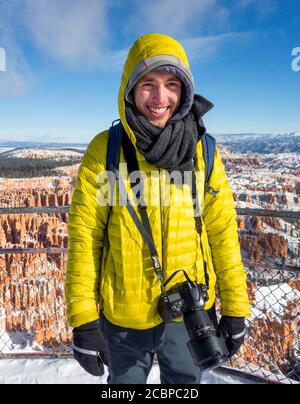Portrait, young man in winter clothes with camera at the viewpoint, rocky landscape with hoodoos in winter, Rim Trail, Bryce Canyon National Park Stock Photo