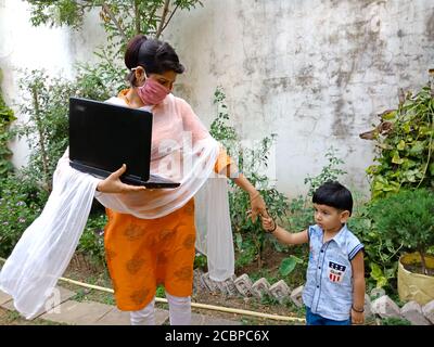 DISTRICT KATNI, INDIA - MAY 16, 2020: An indian girl holing laptop with little kid at garden while wearing face mask. Stock Photo