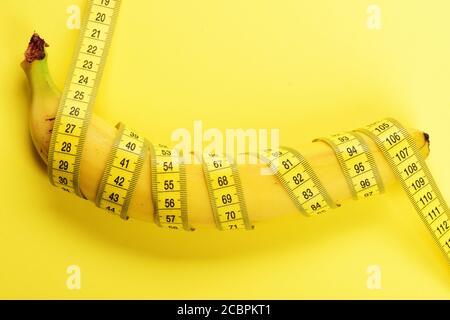 Banana with yellow tape for measuring figure. Rolled centimeter ruler spinned around fresh fruit. Fresh food and healthy diet concept. Yellow tape around banana isolated on yellow background. Stock Photo