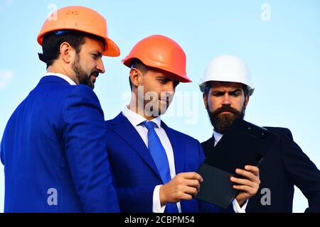 Urban construction and business discussion concept. Board of architects with serious faces in suits and helmets look at tablet. Workers and engineers hold meeting outdoors. Builders discussing budget Stock Photo