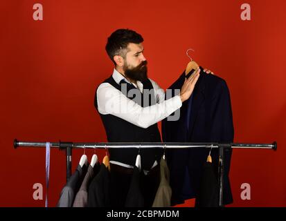 Fashion and individual style concept. Tailor with serious face cleans custom jacket on red background. Man with beard in vest by clothes rack. Designer works near clothes hangers with suits Stock Photo