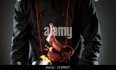 Closeup of chef throwing raw beef steaks from wok pan in fire. Fresh asian food preparation on dark background. Stock Photo