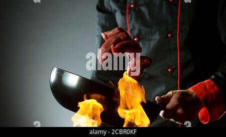 Closeup of chef throwing raw beef steaks from wok pan in fire. Fresh asian food preparation on dark background. Stock Photo