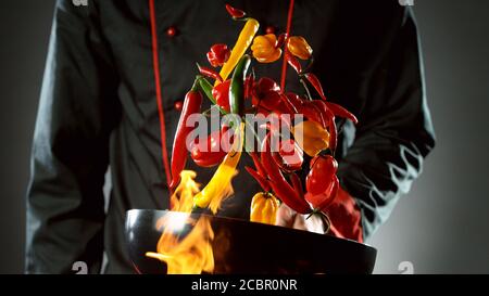 Closeup of chef throwing chilli peppers mix from wok pan in fire. Fresh asian food preparation on dark background. Stock Photo