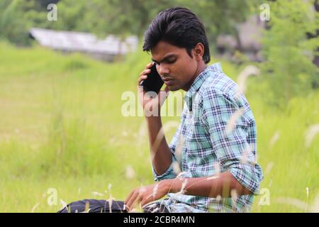 Thoughtful young Asian man talking on mobile phone sitting outside in nature Stock Photo