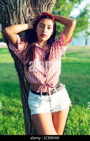 Young Latin girl, long-haired brunette, wearing a shirt of white and brown stripes and red ribbon in her hair, leaning on a tree trunk looking straigh Stock Photo