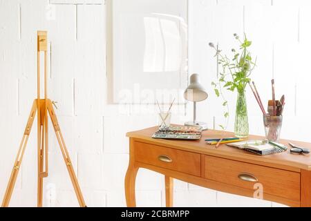 Small space to be creative, crayons, sketchbook, watercolor and brushes on a wooden drawer console and an empty easel against a rough white wall, copy Stock Photo