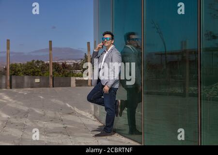 Full body glad male entrepreneur in smart casual outfit smiling and having smartphone conversation while leaning on glass building wall on city street Stock Photo