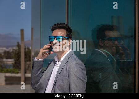 Optimistic adult businessman in sunglasses smiling and talking on smartphone while leaning on glass wall in city Stock Photo