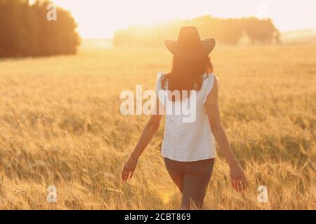Woman farmer in cowboy hat walking with hands on ears at agricultural rye field on sunset Stock Photo