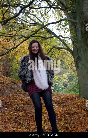 A young lady with long brunette hair laughing and smiling while standing in the golden brown autumn leaves that have fallen to the floor of a forest Stock Photo
