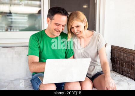 Middle Aged Couple Using Laptop Computer at Home at the Balcony. Couple concept with young people enjoying time together Stock Photo