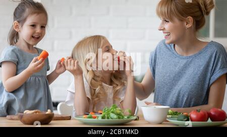 Laughing mother and little daughters playing at kitchen preparing salad Stock Photo