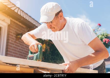 Carpenter old man using drill making holes in a wooden plank or electric saw cutting wood. Woodworking, home improvement, do it yourself (DIY) and con Stock Photo