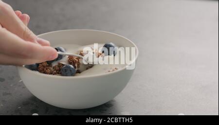 man hand tastes chocolate granola with almond flakes and blueberries in white bowl on concrete surface, wide photo Stock Photo