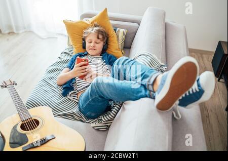 Preteen boy lying with guitar on cozy sofa dressed casual jeans and new sneakers listening to music and chatting using wireless headphones connected w Stock Photo