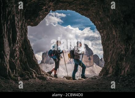 Couple of hikers walking, with Tre Cime di Levarado peaks on background, view from a cave window, in Dolomites, Italy.