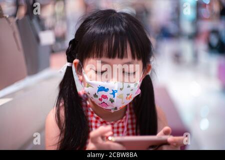 Little child girl playing with smartphone in modern commercial supermarket center with wearing face mask Stock Photo