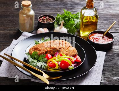 breaded lamb chops served with tomato, red onion, green pepper salad on a black plate on a wooden table with ingredients at the background, landscape Stock Photo