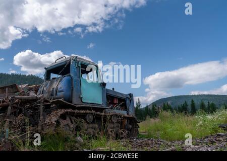 an old abandoned crawler tractor in a field against a cloudy sky, forest and mountains. Stock Photo