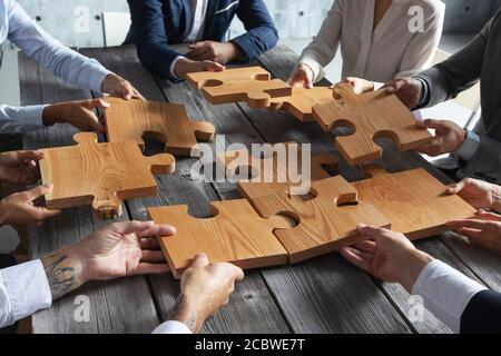 Business people team sitting around meeting table and assembling wooden jigsaw puzzle pieces unity cooperation ideas concept Stock Photo