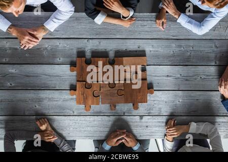 Business people team sitting around meeting table and assembling wooden jigsaw puzzle pieces unity cooperation ideas concept Stock Photo