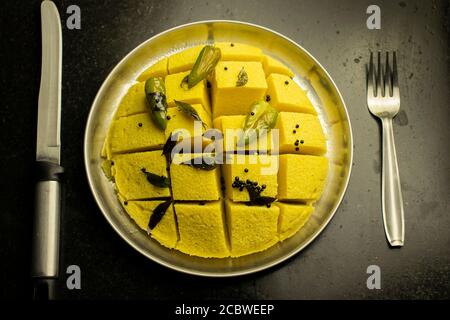 slices of besan dhokla an Indian Gujrati vegetarian food on plate with fork and knife on black background Stock Photo
