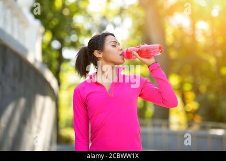 Black girl in pink sweatshirt drinking water during jogging Stock Photo