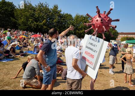 Kiel, Germany. 16th Aug, 2020. A participant of the demonstration 'Freedom and Self-Determination' on the Reventlou meadow holds a banner with the inscription '1.wave? Second wave? permanent wave? Attention demo wave!!' and a model of a corona virus. Organizer of the rally was 'Querdenken Kiel 22'. Credit: Georg Wendt/dpa/Alamy Live News