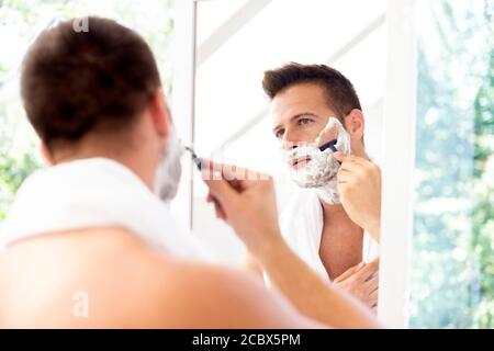 Shot of handsome man with towel around his neck using shaving foam while shawing in front of bathroom mirror. Stock Photo
