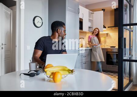 interracial married couple talk during work at home, redhead woman have conversation with african husband sitting with laptop Stock Photo