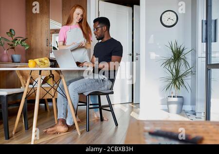interracial married couple talk during work at home, redhead woman have conversation with african husband sitting with laptop Stock Photo