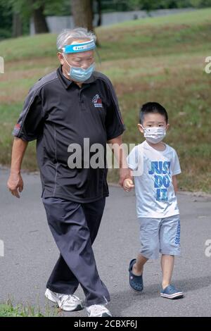 A young Asian American boy walks with his grandfather (presumably) while wearing surgical masks. In a park in Flushing, Queens, New York City. Stock Photo