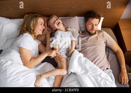 mother and daughter have fun on bed while father is sleeping, family on bed, weekends concept Stock Photo
