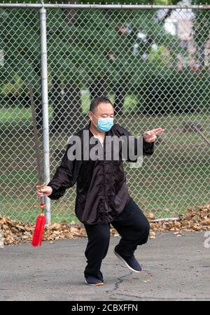 An Asian American man wearing a face mask leads a small Tai Chi class holding a sword. In a park in Queens, New York City. Stock Photo