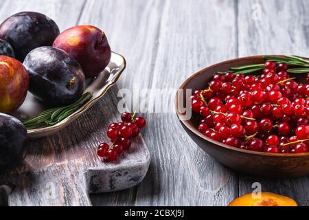 Fresh sweet plum fruits whole and sliced in plate with rosemary leaves on old cutting board with red currant berries in wooden bowl Stock Photo