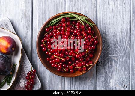 Fresh red currant berries in wooden bowl near to sweet plum fruits in plate with rosemary leaves on old cutting board, grey wood background, top view Stock Photo