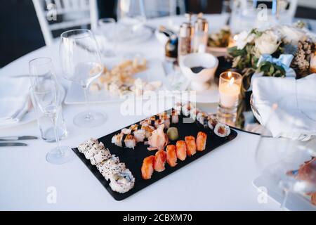 Close-up of a black plate for serving sushi and rolls on a white tablecloth on a table with glasses and plates on a blurred background. Stock Photo