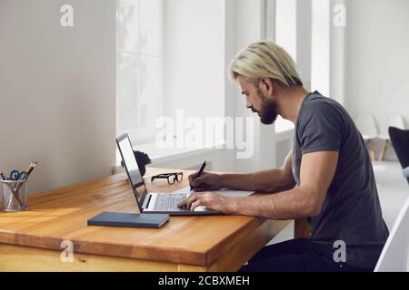 Young guy writing down main points from online business conference or educational webinar at desk indoors Stock Photo
