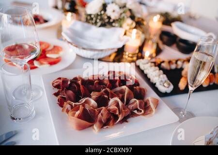 Close-up of a square white plate with prosciutto on a festively served table with sushi set and tomatoes on a white tablecloth. Stock Photo