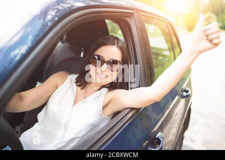 A smart looking middle-aged adult woman wearing sunglasses put her hand out the car window and raised her thumbs up. Stock Photo