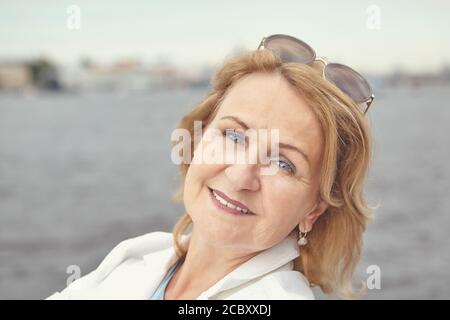 Beautiful active caucasian senior woman about 62 years old with blond hair and sunglasses is posing for photo near river in Saint-Petersburg and smili Stock Photo