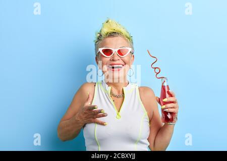Cheerful elderly lady with short yellow hair in funny outfit drinking red cocktail and laughing. close up portrait, studio shot.lifestyle, happiness, Stock Photo