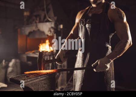 muscular forger man with beard using hammer for beating iron manufacture at smithy, wearing leather apron or uniform Stock Photo