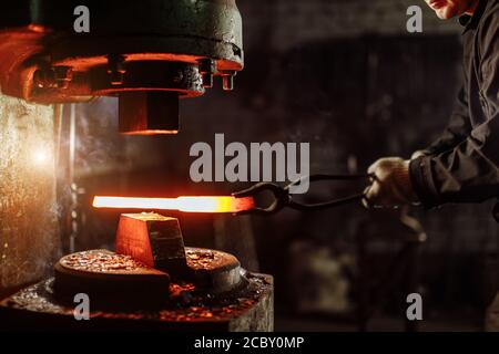 muscular forger man with beard using hydraulic press for steel arms manufacture at smithy, wearing leather apron or uniform Stock Photo