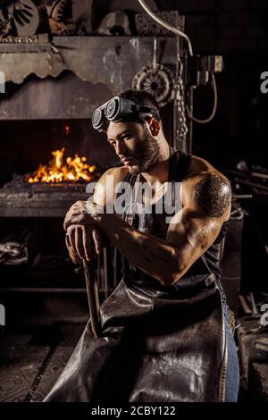 portrait of thoughtful muscular blacksmith holding hammer and looking down, wearing leather uniform. furnace in the background Stock Photo