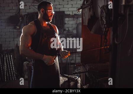 male blacksmith forging in his workshop, make iron products for manifacture of fireplaces and stoves. Use furnace for heating metal. Wearing protectiv Stock Photo