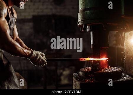 muscular forger man with beard using hydraulic press for steel arms manufacture at smithy, wearing leather apron or uniform Stock Photo