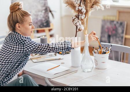 beautiful blond girl with bun in stylish casual clothes choosing a pencil for drawing. close up side view photo. copy space. Stock Photo