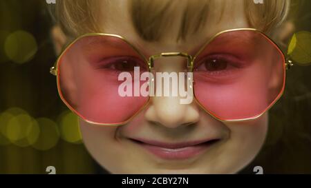 Close up face footage of child. Smiling, looking at camera. Little fun blonde kid teen teenager girl 4-5 years old in pink sunglasses posing isolated on golden dark background in studio. 4k footage Stock Photo